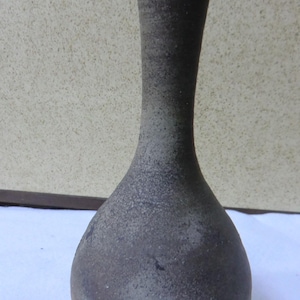 以下が含まれることがあります： 手作りの、暗い灰色のセラミック花瓶。表面はざらざらとした質感。丸みを帯びた底、細い首、広がった開口部が特徴。全体的な形状は涙のしずくを連想させる。花を飾ったり、装飾品として使用するのに適した花瓶です。