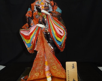 Pode incluir: Uma boneca japonesa Ryukyu, apresentando uma mulher em um quimono tradicional com padrões florais vibrantes em laranja, preto e azul. A boneca tem uma expressão serena e é exibida em uma base preta com uma plataforma dourada. Uma pequena placa de madeira está incluída.