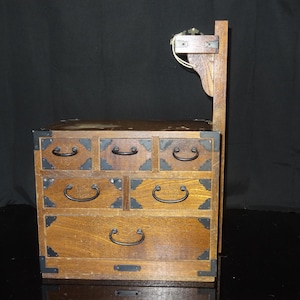 Puede incluir: Una caja de joyería de madera con seis cajones y un soporte decorativo de madera. La caja está hecha de madera de color marrón claro con esquinas y tiradores de cajón de metal negro. La parte superior de la caja tiene un acabado marrón oscuro.