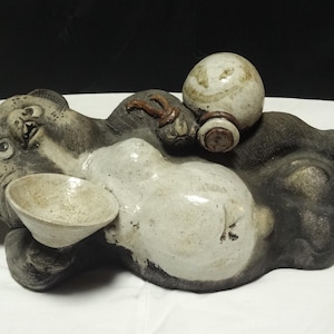May include: A ceramic tanuki figurine in a reclining position, featuring a dark brown and cream-colored glaze. The figure holds a small white bowl in one hand and a round, light-colored sake bottle in the other, with a textured, weathered finish.