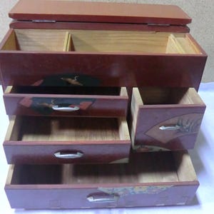 May include: A red wooden jewelry box with five drawers. The drawers have intricate designs and are decorated with gold accents. The top drawer is open and reveals a small compartment.
