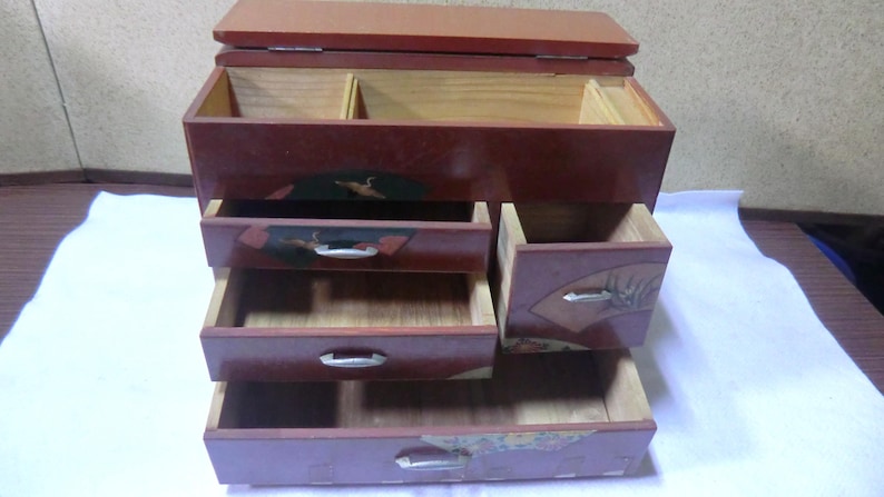 May include: A red wooden jewelry box with five drawers. The drawers have intricate designs and are decorated with gold accents. The top drawer is open and reveals a small compartment.