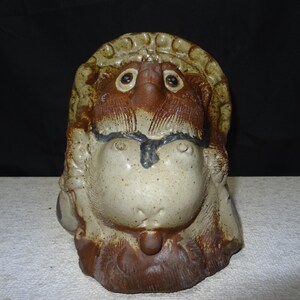 May include: A ceramic sculpture of a stylized animal head with large eyes and a neutral expression. The sculpture features a mix of brown, beige, and black colors, with a textured surface. The piece has a unique, handcrafted appearance.