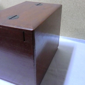 May include: A dark brown wooden box with a hinged lid and two metal clasps. The box has a small slot on the front side.