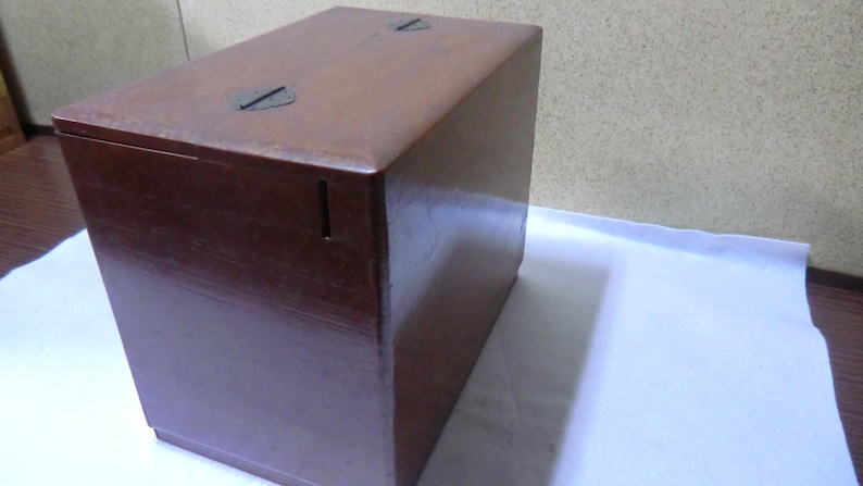 May include: A dark brown wooden box with a hinged lid and two metal clasps. The box has a small slot on the front side.