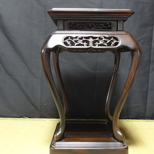 May include: A dark brown wooden display stand featuring a square top, four curved legs, and intricate openwork carvings along the upper apron. The stand has a smooth, polished finish and a square base, providing a stable platform for decorative items.