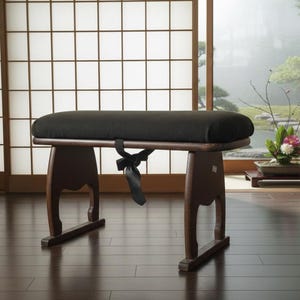 Puede incluir: Un banco de madera marrón oscuro con un cojín negro. El banco tiene patas curvas y una correa negra atada alrededor del marco. El fondo presenta una habitación de estilo japonés con una pantalla shoji y una vista de un jardín.