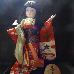 Puede incluir: Muñeca japonesa con kimono tradicional. La muñeca tiene cabello negro y lleva un kimono rojo con estampados florales y una faja color crema. La muñeca se exhibe sobre una superficie negra con una placa de madera.