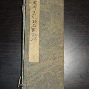 Op de afbeelding: Een vintage boek met een vervaagde grijze kaft en een verticaal papieren label met Japanse tekst. Het boek is gebonden met een metalen sluiting en lijkt verouderd. De kaft heeft een textuurpatroon.