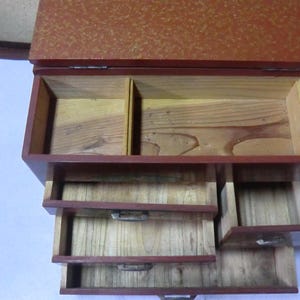 May include: A red wooden box with a hinged lid and four drawers. The box is made of wood with a red finish and has a decorative pattern on the lid. The box has four drawers that are all open and empty.