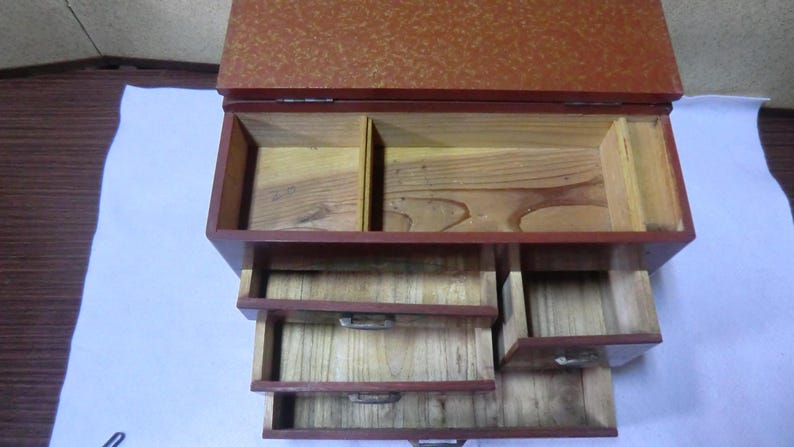 May include: A red wooden box with a hinged lid and four drawers. The box is made of wood with a red finish and has a decorative pattern on the lid. The box has four drawers that are all open and empty.