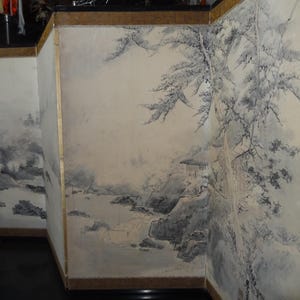 May include: A multi-panel screen featuring a traditional Asian landscape painting. The artwork depicts mountains, water, and trees in shades of gray and white, with a hint of gold trim on the wooden frame.