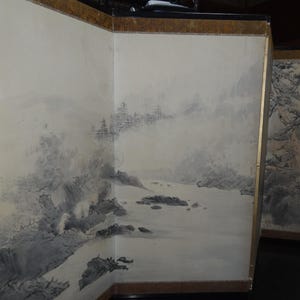 May include: A three-panel folding screen with a landscape painting. The artwork depicts a misty mountain scene with a river and trees. The screen has a gold-colored border.