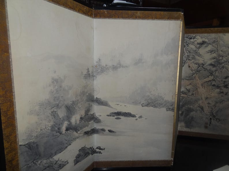 May include: A three-panel folding screen with a landscape painting. The artwork depicts a misty mountain scene with a river and trees. The screen has a gold-colored border.