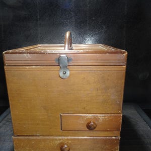 Puede incluir: Una caja de madera vintage con asa, cierre metálico y dos pequeños cajones con pomos redondos. La caja tiene un acabado marrón claro con desgaste visible, lo que sugiere edad y uso. El asa es un lazo de metal curvado.