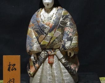 May include: A Japanese doll with a white face and black hair, wearing a multi-colored patterned kimono with gold, grey, and pink floral details. The doll stands on a black rectangular base next to a small wooden plaque with black Japanese text.