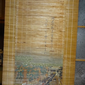May include: A vertical bamboo scroll featuring a painted scene of a procession leading to a shrine, with Japanese calligraphy text above the illustration and at the bottom. The scroll is constructed from thin, light brown bamboo slats.