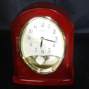 May include: A Citizen brand, red wooden mantel clock with a gold-tone oval clock face. The clock face has black numerals and hands, and a white pendulum. The clock is a classic design.