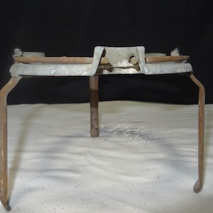 May include: A vintage metal tripod stand with a circular top and three legs. The stand is made of weathered metal with a rusty appearance, suggesting age and use. The background is a plain black.