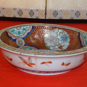 Puede incluir: Un cuenco ovalado de cerámica antigua con exterior blanco e interior detallado. El interior presenta un fondo rojo y dorado con motivos florales y geométricos en azul, verde y blanco. El exterior tiene diseños de mariposas e insectos naranjas.