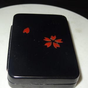 Puede incluir: Una caja rectangular negra con un acabado brillante. La caja está decorada con una flor de cerezo roja y un solo pétalo. La caja está cerrada y parece estar hecha de un material duro.