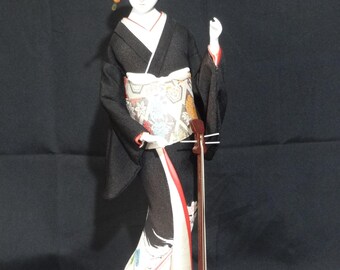 May include: A Japanese geisha doll in a black kimono with a white face and elaborate hairstyle. The doll holds a traditional shamisen instrument. The kimono features colorful floral patterns and a red lining. The doll stands on a small wooden base.