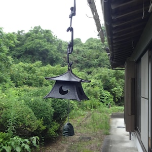 以下が含まれることがあります： 三日月型の切り抜きが施された塔のような屋根を持つ黒い金属製の風鈴。小さな猿のフィギュアが連なった鎖で吊るされています。塔の下には小さな鐘がぶら下がっており、白い山のイラストが描かれた青い短冊が取り付けられています。