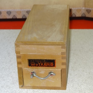 Op de afbeelding: Een rechthoekige houten kist met een lade en een zilveren handvat. De kist heeft een rechthoekig label met Japanse tekst. De lade is gesloten.