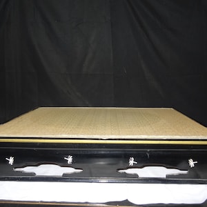 Puede incluir: Una tabla rectangular de madera lacada en negro con una superficie de paja tejida. La base presenta recortes decorativos y pequeños motivos florales pintados en blanco en el borde frontal, con un fino borde dorado en la parte superior.