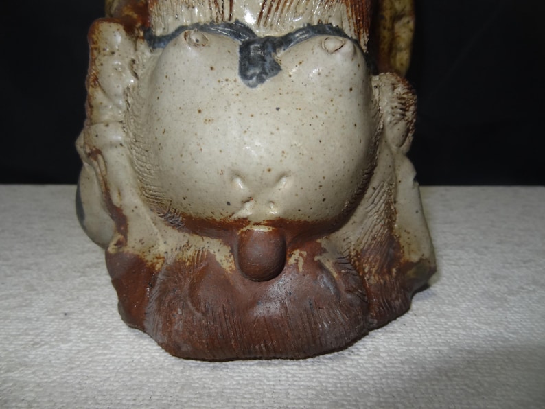 May include: A ceramic sculpture of a stylized animal head, featuring a light beige face with dark brown spots and a dark brown lower section. The sculpture has a textured surface and a small, rounded protrusion.
