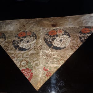 Op de afbeelding: Driehoekig stuk stof met een bloemenpatroon. De stof heeft een gouden achtergrond met grote cirkelvormige ontwerpen met witte en zwarte bloemen met oranje accenten. De rand is een lichter goud met een bloemenpatroon.