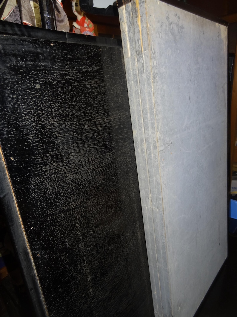 May include: A stack of gray panels with visible wood grain and a black panel with a textured surface. The panels are leaning against each other. The image also shows a glimpse of a traditional Japanese doll.