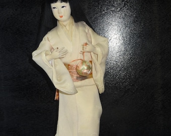 Puede incluir: Muñeca japonesa con kimono crema con estampado floral y obi rosa. La muñeca tiene cabello negro, rostro pálido y sostiene un pequeño adorno redondo. La muñeca está sobre una superficie negra.