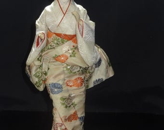 Pode incluir: Boneca japonesa em quimono tradicional. A boneca tem cabelo preto e rosto pálido. O quimono é creme com estampas florais em laranja, azul e verde. O obi é laranja e o forro vermelho. A boneca está sobre uma base retangular.