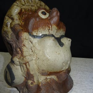 May include: A ceramic sculpture of a creature's head, featuring a tan and brown color palette. The sculpture has a textured surface with detailed facial features, including a prominent nose, eye, and mouth. The overall design suggests a whimsical or mythological theme.