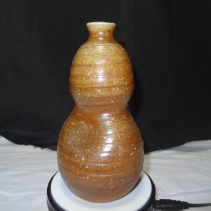 May include: A handmade, gourd-shaped ceramic vase with a speckled, brown glaze. The vase has a narrow neck and a rounded body, with horizontal textured lines. The pottery is displayed on a white surface against a black backdrop.