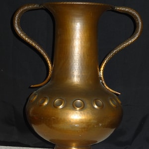 以下が含まれることがあります： 2 つの湾曲したハンドルが付いた、背の高いアンティーク銅の花瓶。花瓶は、ベースの上部近くに一連の円形の切り欠きがある球根状のボディを持っています。銅は暖かく、古くなった緑青を持っています。