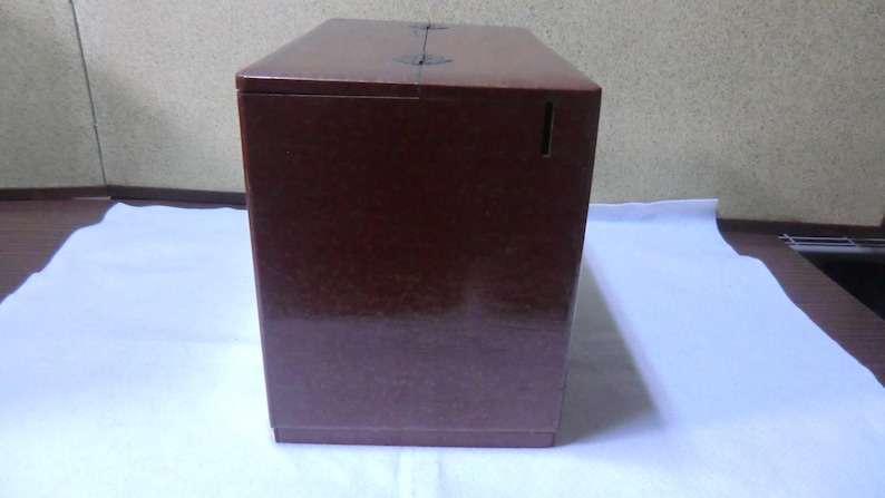 May include: A dark brown wooden box with a hinged lid and a small slot on the front. The box has a textured surface and appears to be made of a high-quality material.