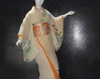 Puede incluir: Una muñeca japonesa geisha con un kimono tradicional. La muñeca lleva un kimono color crema con detalles en naranja y un obi decorativo. El kimono está adornado con estampados florales y paisajísticos. La muñeca tiene el pelo negro y posa con un brazo extendido.