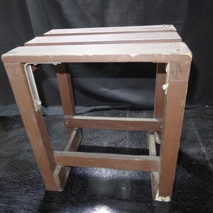以下が含まれることがあります： 長方形の天板と4本の脚が付いた茶色の木製ステップスツール。 スツールは、ネジと金属製のブラケットが見えるシンプルなデザインです。 木材には摩耗の兆候が見られ、中古品であることを示唆しています。