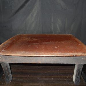 May include: A small, rectangular wooden table with a dark brown tabletop and black legs. The tabletop has a slightly distressed appearance, with visible wood grain and a warm, reddish-brown hue. The legs are simple and sturdy.