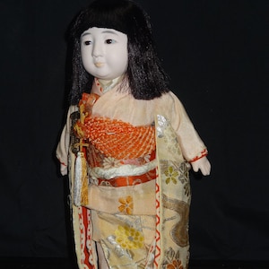 Pode incluir: Boneca japonesa com cabelo preto e tez pálida. A boneca usa um quimono creme com detalhes em laranja e dourado. O quimono apresenta padrões florais e uma faixa decorativa.