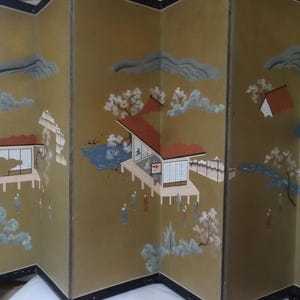 May include: A four-panel folding screen featuring a gold-toned background with painted illustrations of traditional buildings, stylized trees, and small figures. The artwork includes blue clouds, red-roofed structures, and white flowering branches.
