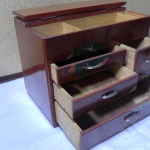 May include: A small, wooden chest with a hinged lid and four drawers. The chest is painted red and has intricate designs on the drawers. The chest is open, revealing the empty drawers.