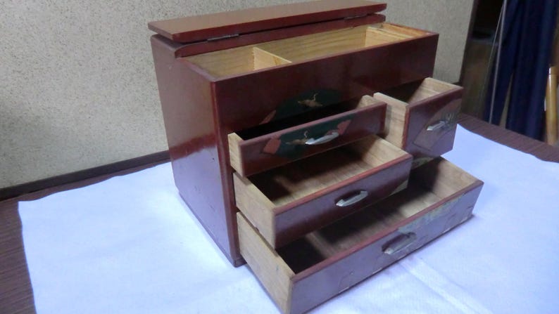 May include: A small, wooden chest with a hinged lid and four drawers. The chest is painted red and has intricate designs on the drawers. The chest is open, revealing the empty drawers.