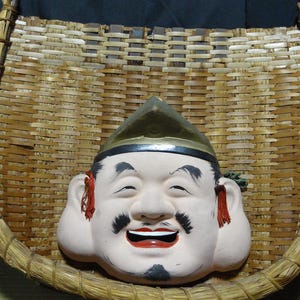 May include: A woven bamboo basket with a curved, U-shaped frame featuring a central ceramic mask of a smiling figure. The mask has a pale complexion, black painted eyebrows and facial hair, red lips, and a gold-colored pointed headpiece.
