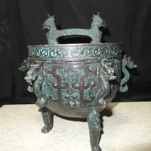 Puede incluir: Quemador de incienso de bronce con tres patas, adornado con dragones y diseños geométricos. El asa presenta dos figuras de dragón. El bronce tiene una pátina verde y marrón. Altura aproximada de 20 cm.
