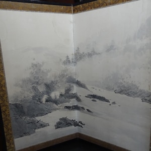 May include: A two-panel folding screen with a landscape painting. The artwork features a monochromatic scene of mountains, water, and trees in shades of gray and black. The screen has a gold-colored border.