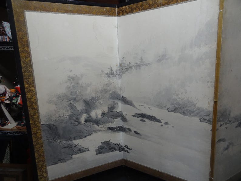 May include: A two-panel folding screen with a landscape painting. The artwork features a monochromatic scene of mountains, water, and trees in shades of gray and black. The screen has a gold-colored border.