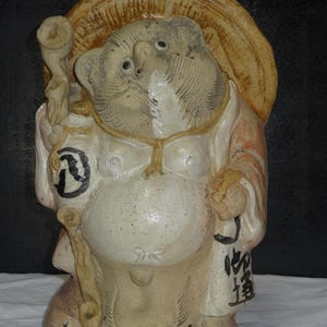 Peut inclure: Statue en céramique de Tanuki, une figure traditionnelle japonaise de chien viverrin. La statue est beige et crème, avec un chapeau de paille et un bâton. Elle a un gros ventre et tient un sac avec des caractères japonais.